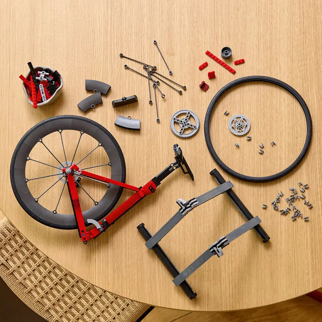 LEGO Icons Road Bike build in red, black and gray plastic bricks is partly assembled beside wheel, spoke, gear and stand components arranged across a light wood table.
