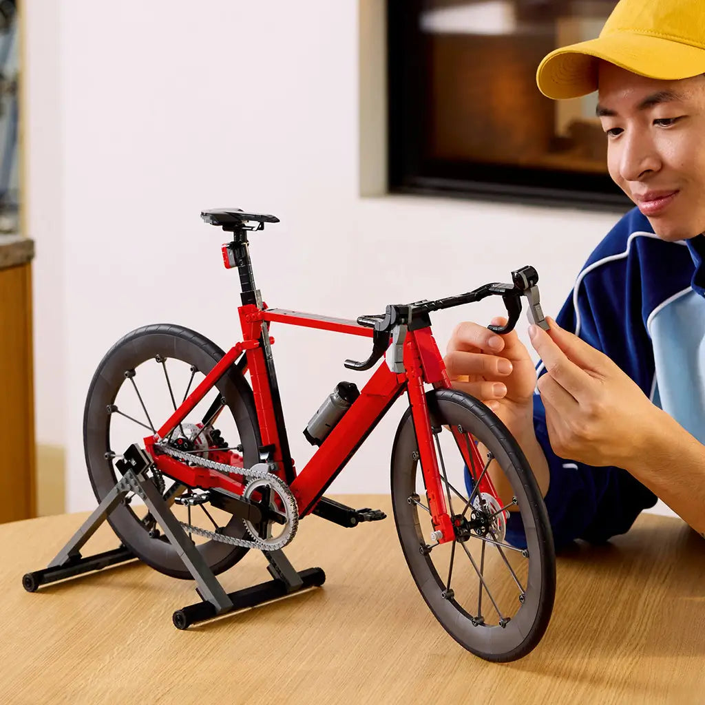 LEGO Icons Road Bike model in red and black bricks sits on a gray stand as a person adjusts the drop handlebar at a wooden table in a softly lit home interior.