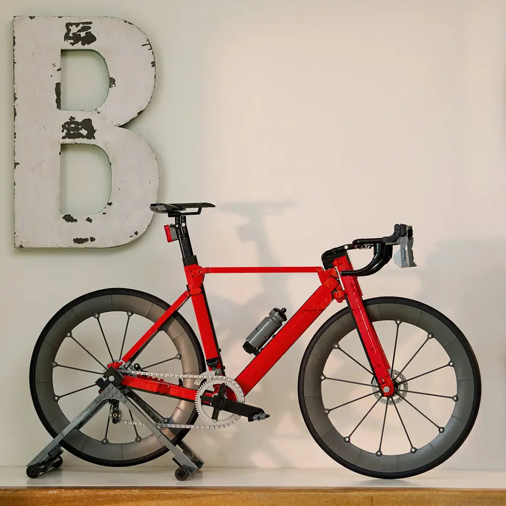 LEGO Icons Road Bike display model in red and black plastic bricks rests on a gray stand atop a wooden shelf, beneath a distressed white letter B on a pale wall.