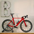 LEGO Icons Road Bike display model in red and black plastic bricks rests on a gray stand atop a wooden shelf, beneath a distressed white letter B on a pale wall.