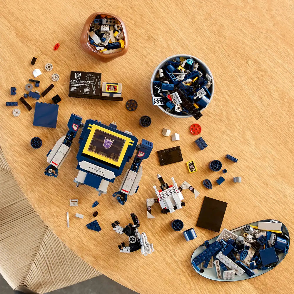LEGO Icons Soundwave 10358 build in progress with scattered blue and gray LEGO pieces, showcasing partially built robot, cassette builds, and organized components on a wooden tabletop.