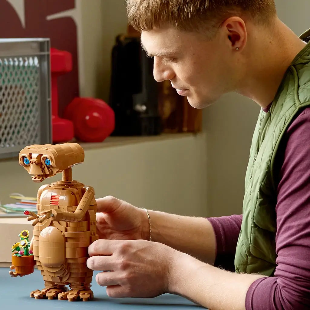 LEGO Ideas E.T. model in warm brown bricks with blue eyes is adjusted by a builder at a table, with the alien holding a flowerpot and a glowing red fingertip.