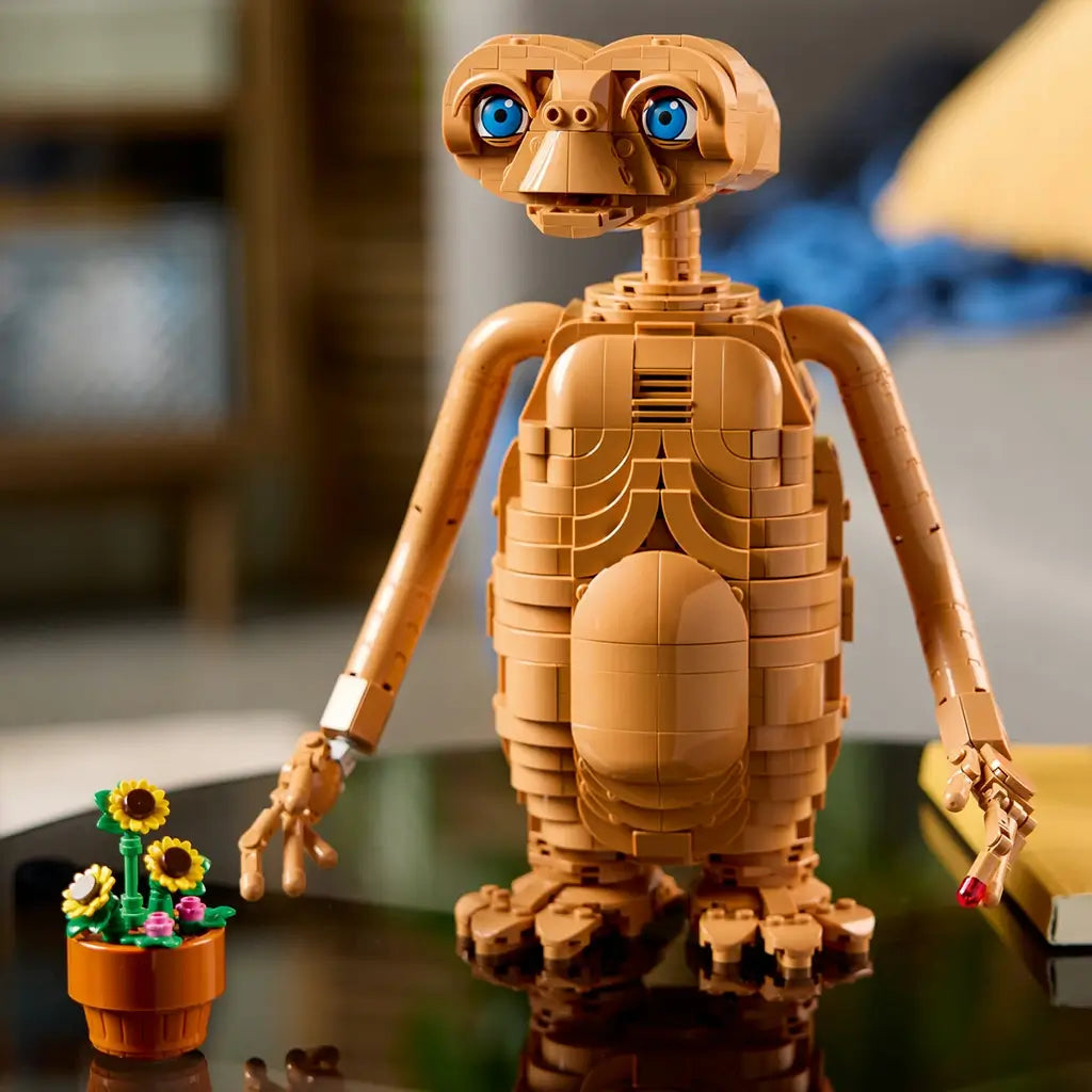 LEGO Ideas E.T. model in warm brown bricks features bright blue eyes, long arms and detailed feet, posed with a flowerpot of sunflowers on a glossy tabletop.