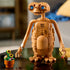 LEGO Ideas E.T. model in warm brown bricks features bright blue eyes, long arms and detailed feet, posed with a flowerpot of sunflowers on a glossy tabletop.
