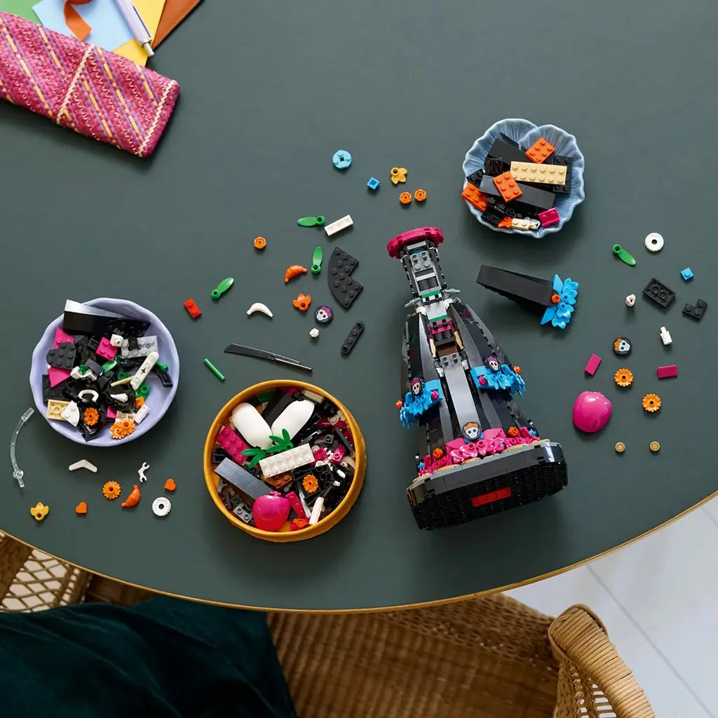 LEGO Ideas La Catrina building set in black, magenta, blue and orange bricks lies partly assembled on a dark tabletop, surrounded by bowls of pieces and floral details.