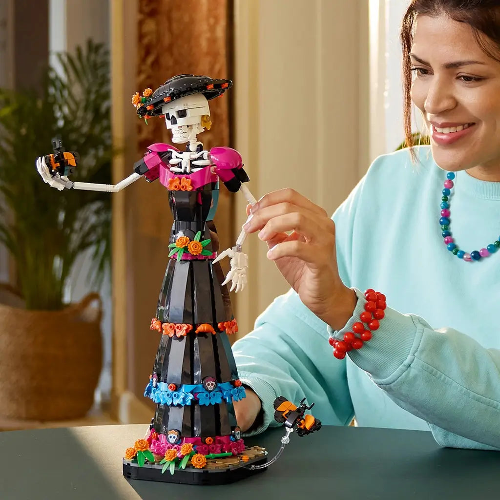 LEGO Ideas La Catrina model in black, white, magenta and orange bricks is adjusted by a smiling builder, with a butterfly accessory displayed beside the floral dress.
