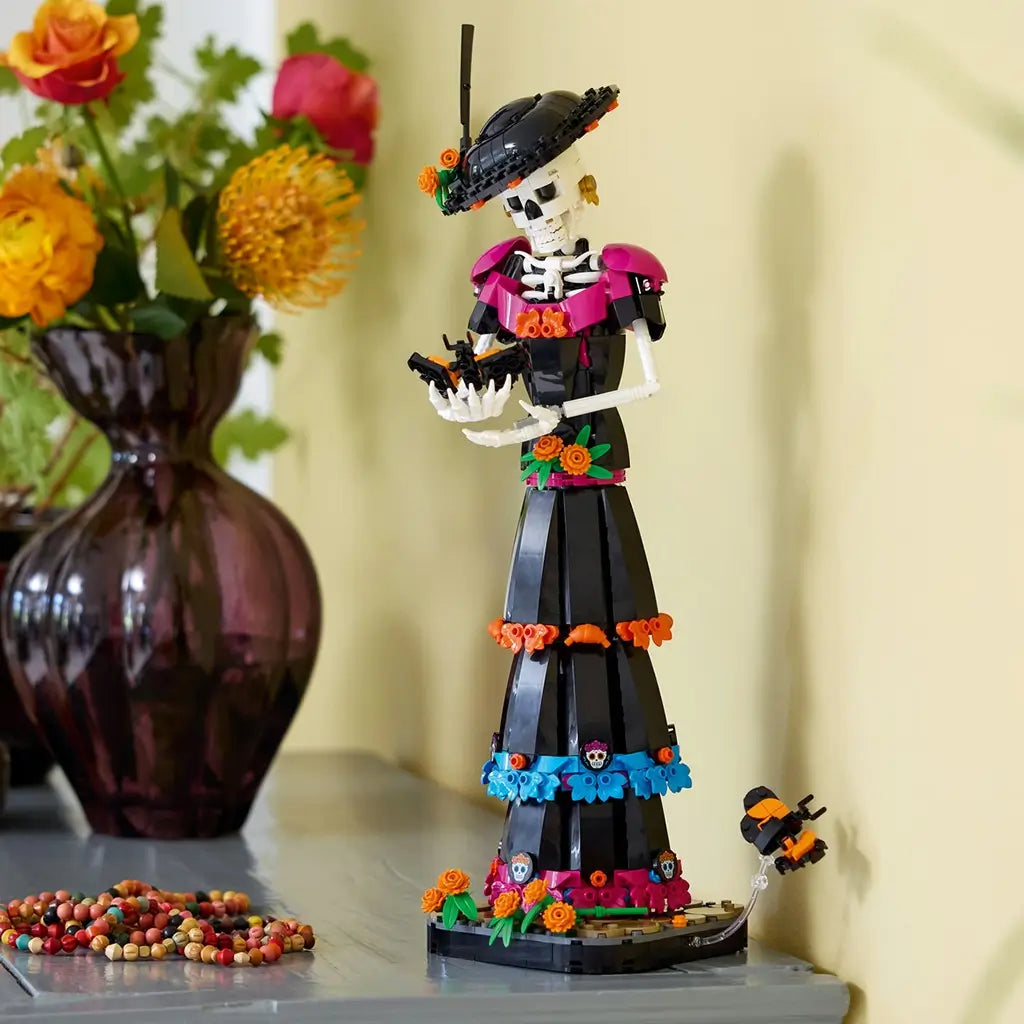 LEGO Ideas La Catrina display model features a white skeleton in a black floral dress with magenta, blue and orange accents, posed beside a vase of flowers on a mantel.