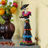 LEGO Ideas La Catrina display model features a white skeleton in a black floral dress with magenta, blue and orange accents, posed beside a vase of flowers on a mantel.