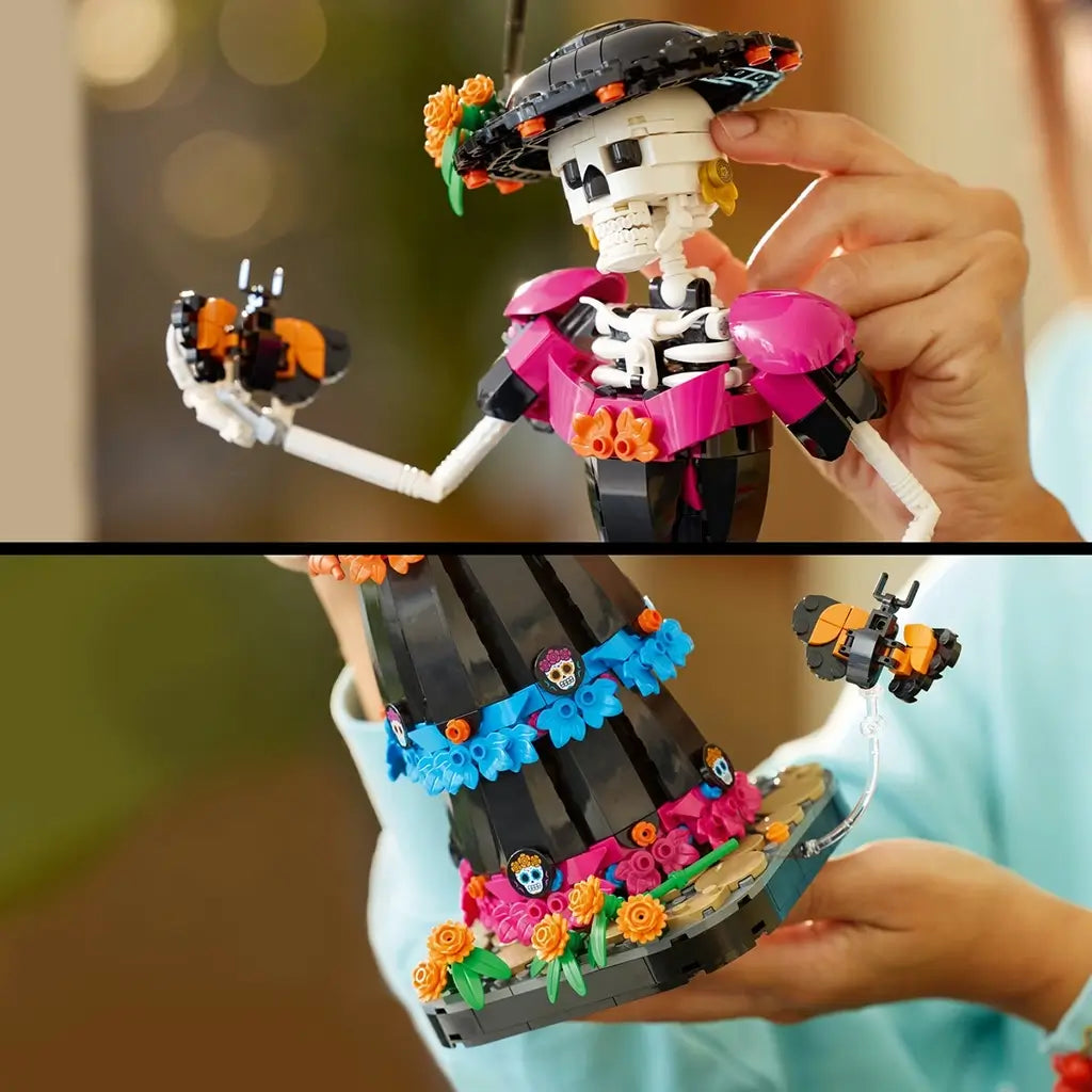 LEGO Ideas La Catrina model features a white skeleton in a black dress with magenta, blue and orange floral details, holding a butterfly while hands adjust her hat.