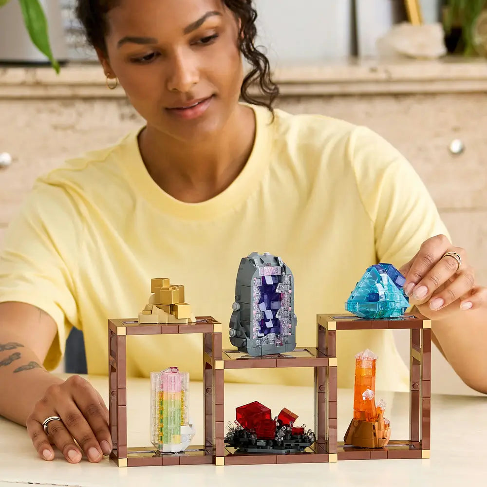 A person wearing a yellow shirt builds the LEGO Ideas Mineral Collection set, arranging translucent crystal structures in shades of blue, red, purple, and gold on wooden-style frames.