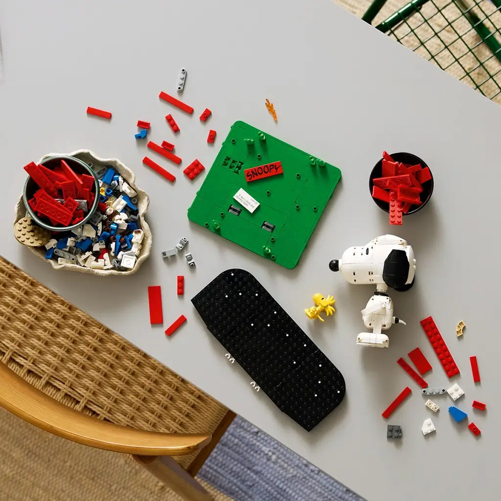 LEGO Ideas Snoopy's Doghouse parts in red, white, green and black bricks surround a partly built base, Snoopy figure and yellow Woodstock on a gray tabletop.