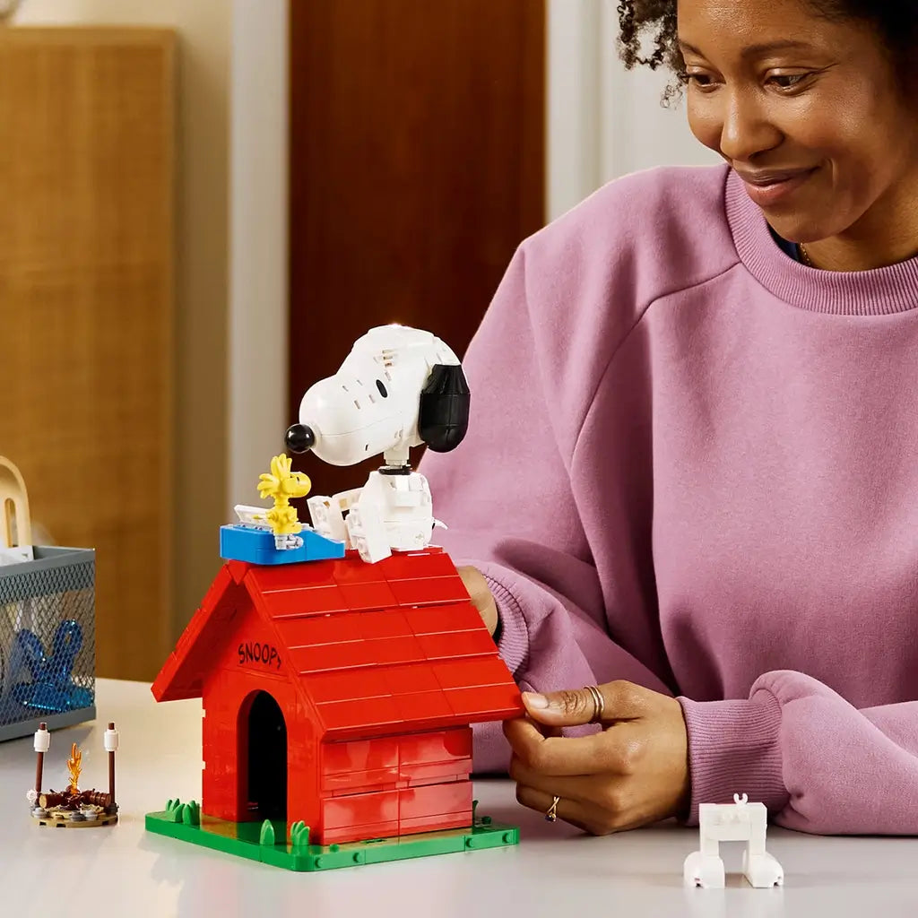 LEGO Ideas Snoopy's Doghouse in red, white and green bricks is handled by a builder at a table, with Snoopy and yellow Woodstock posed beside a blue typewriter on the roof.