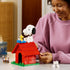LEGO Ideas Snoopy's Doghouse in red, white and green bricks is handled by a builder at a table, with Snoopy and yellow Woodstock posed beside a blue typewriter on the roof.