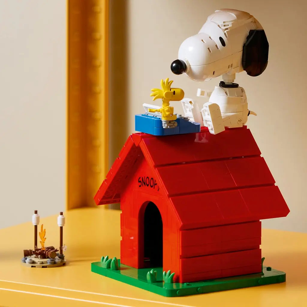 LEGO Ideas Snoopy's Doghouse in red and green bricks features white Snoopy and yellow Woodstock beside a blue typewriter on the roof, displayed with a small campfire.