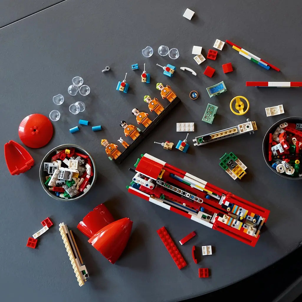 LEGO Ideas Tintin Moon Rocket building set shows red, white and multicolor plastic bricks, minifigures and components arranged for assembly across a dark tabletop workspace background.