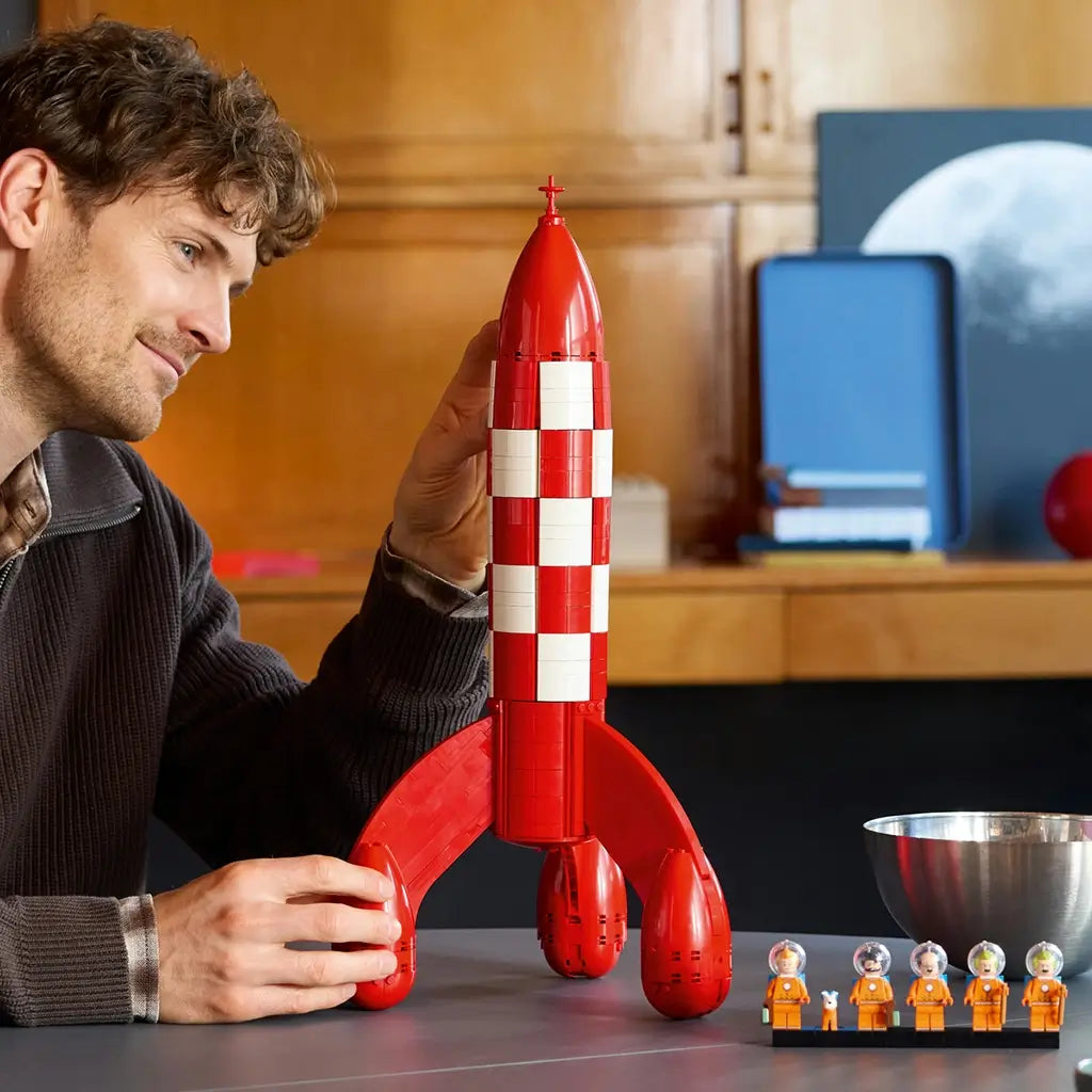LEGO Ideas Tintin Moon Rocket display model features a red and white plastic rocket with curved fins as an adult builder adjusts the structure beside astronaut minifigures indoors.