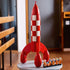 LEGO Ideas Tintin Moon Rocket model features a red and white plastic rocket with curved fins and astronaut minifigures, standing on a tabletop in a warmly lit indoor room setting.