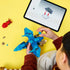 LEGO Jurassic World Baby Dinosaur Pteranodon set shows a child assembling the blue and tan brick-built flying dinosaur beside a tablet with digital instructions on yellow.