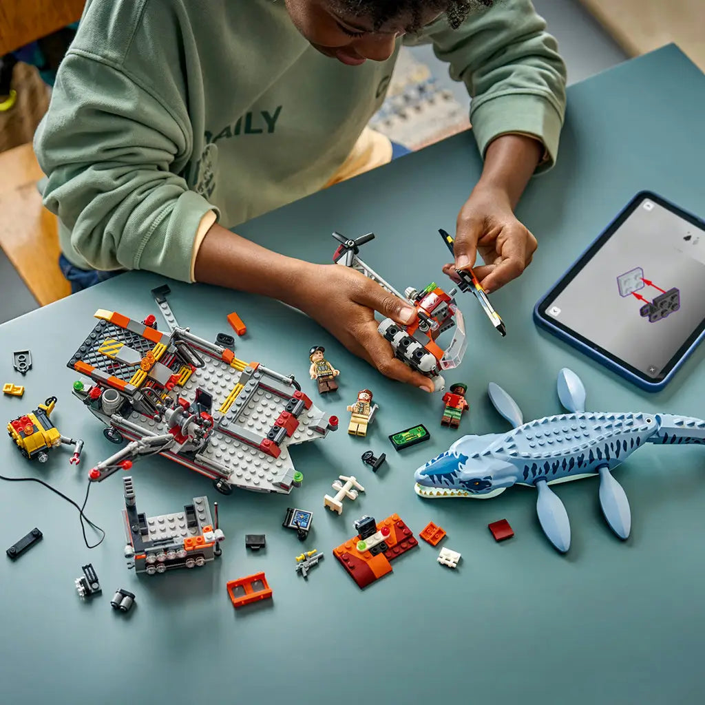 LEGO Jurassic World Mosasaurus building set fills a teal tabletop with the blue-gray dinosaur, partly built rescue boat, helicopter, minifigures and loose plastic bricks.