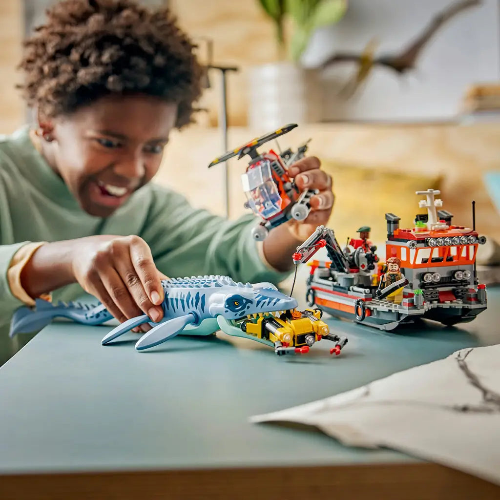 LEGO Jurassic World Mosasaurus playset features a blue-gray marine reptile facing a yellow submersible as a child holds the red helicopter beside the rescue boat.