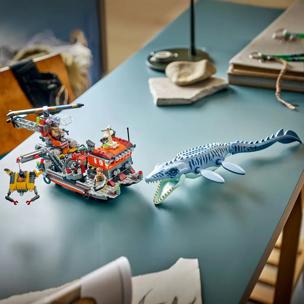 LEGO Jurassic World Mosasaurus playset shows the blue-gray marine reptile with open jaws beside an orange rescue boat, red helicopter and yellow submersible on a teal table.