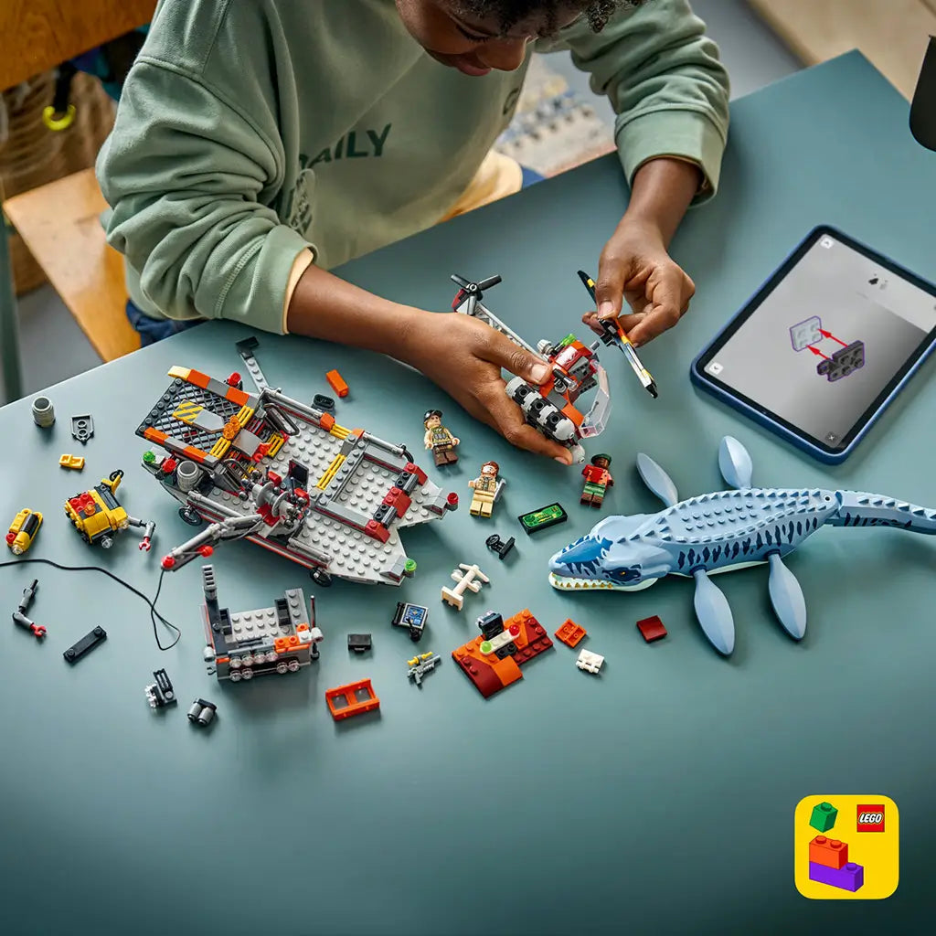 LEGO Jurassic World Mosasaurus building set is spread across a teal table as a child assembles the red helicopter beside the blue-gray dinosaur, boat and loose bricks.