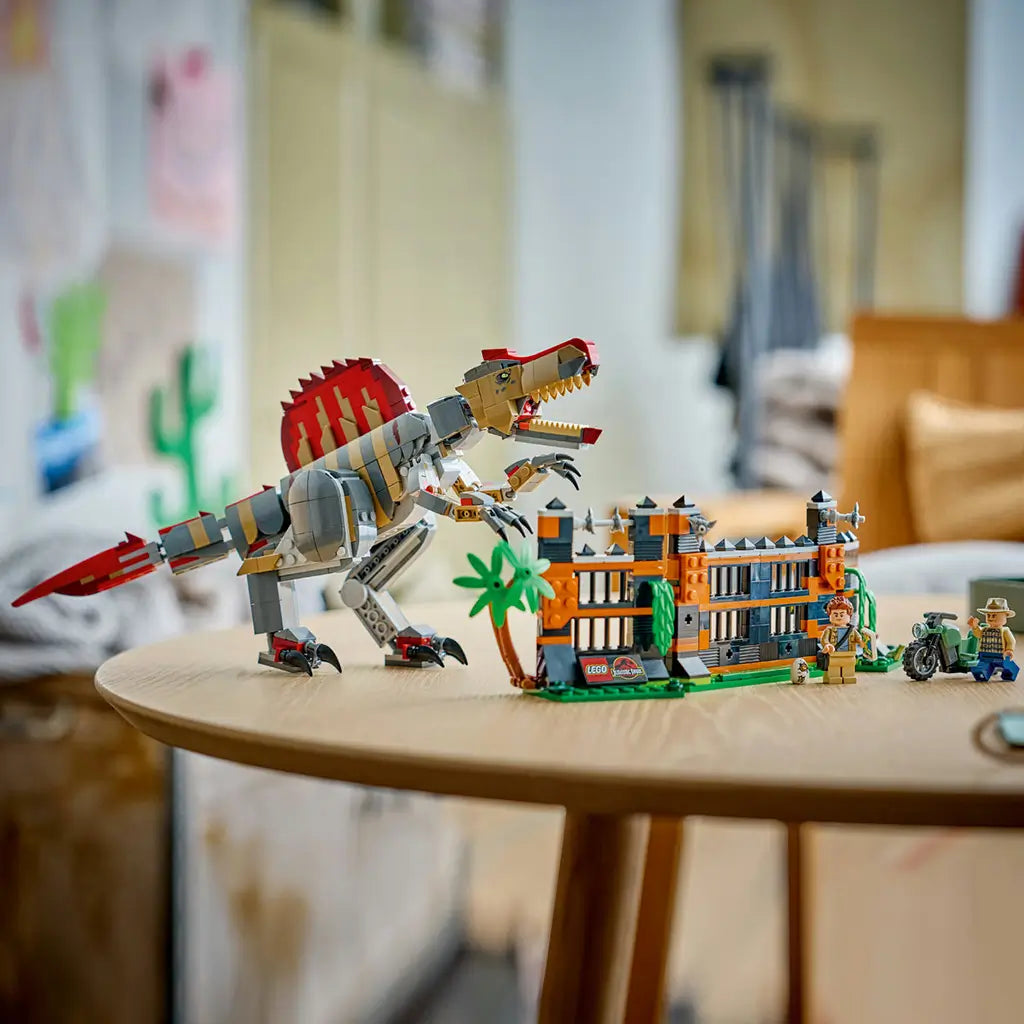 LEGO Jurassic World Spinosaurus set displays a gray, tan and red brick dinosaur beside an orange and black enclosure, green motorcycle and minifigures on a wood table.