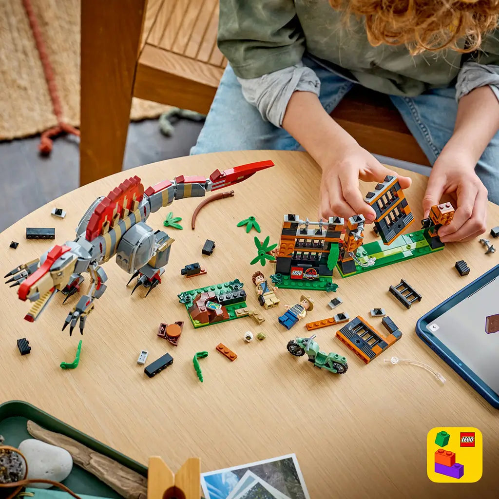 LEGO Jurassic World Spinosaurus building set covers a wood table with a gray and red dinosaur, orange and black enclosure pieces, minifigures and loose plastic bricks.