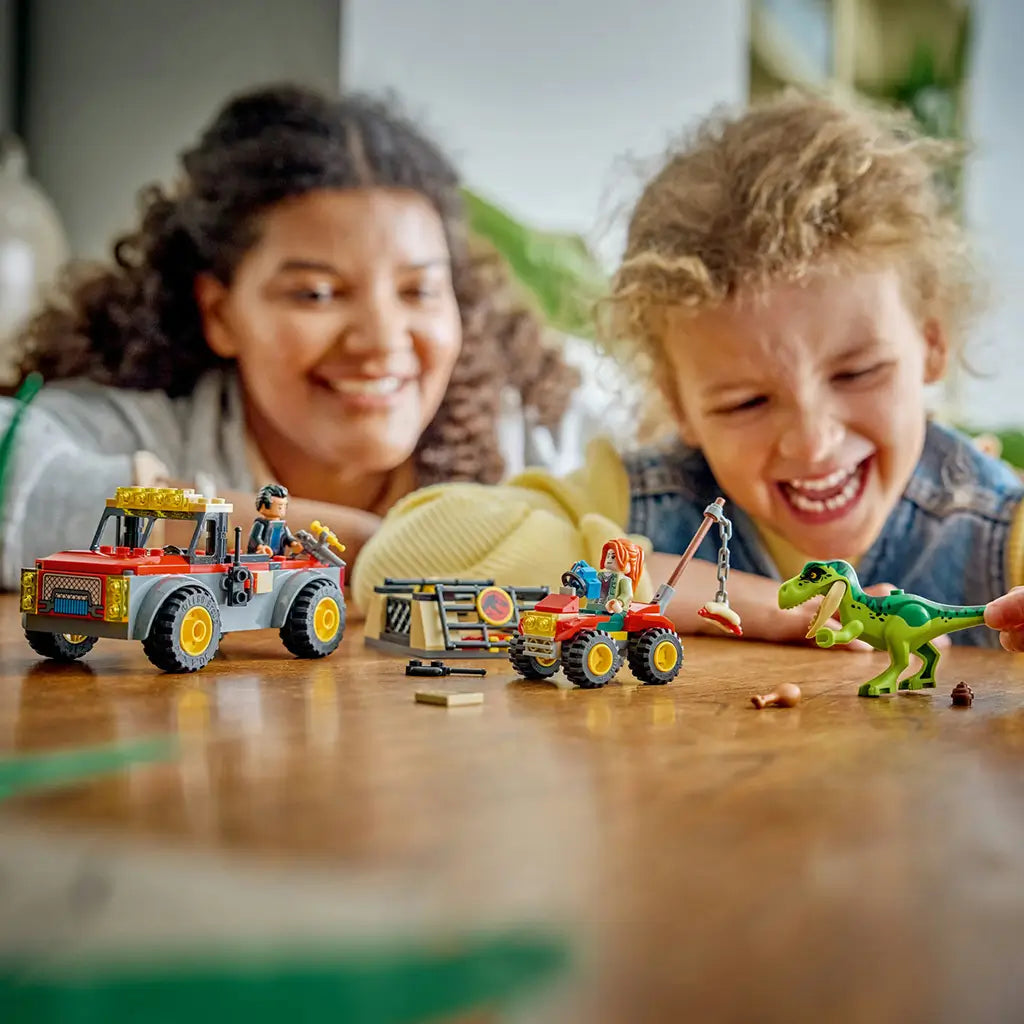 LEGO Jurassic World Young T. rex playset shows a green dinosaur, red and gray transport truck and red ATV with two minifigures as a child plays at a wooden table.