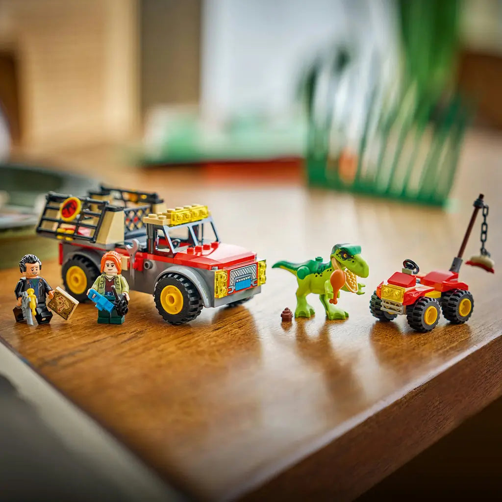 LEGO Jurassic World Young T. rex set displays a green dinosaur with food bait beside a red and gray transport truck, red ATV and two minifigures on a wooden table.