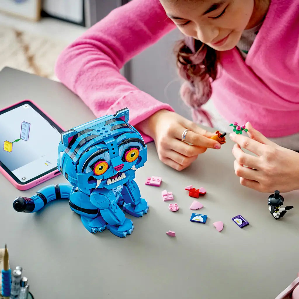 LEGO KPop Demon Hunters building set uses bright blue plastic bricks as a child assembles accessories beside the Derpy Tiger model, Sussie Bird and a digital tablet.