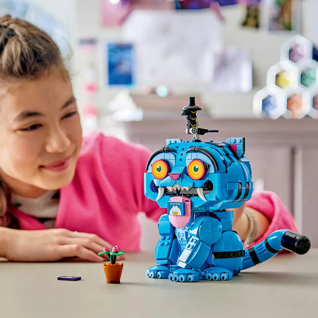 LEGO KPop Demon Hunters Derpy Tiger toy uses bright blue plastic bricks with orange eyes and white fangs as a child plays with the model and flower pot on a tabletop.