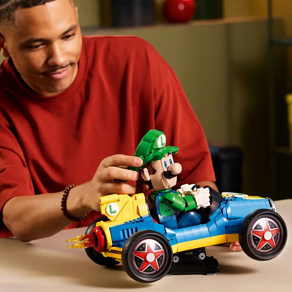 LEGO Mario Kart Luigi model features green, blue and yellow plastic bricks, a detailed character figure seated in a kart, being adjusted by a builder at a tabletop to demonstrate display features.