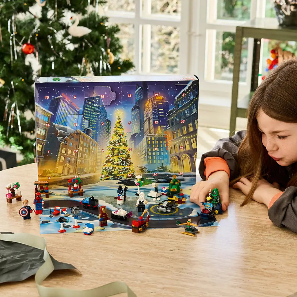 LEGO Marvel Advent Calendar 2026 play scene shows a child arranging colorful superhero minifigures and mini builds before a snowy city calendar near a decorated Christmas tree.
