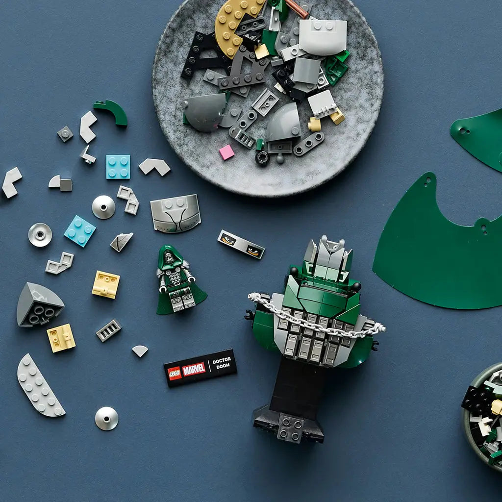 LEGO Marvel Doctor Doom Bust pieces in dark green, silver, gray and black surround a partly assembled model and minifigure on a blue work surface during building.