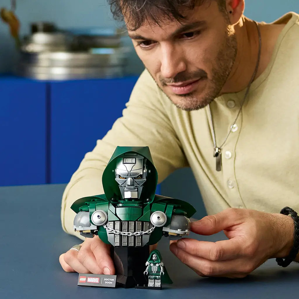 LEGO Marvel Doctor Doom Bust building set is assembled by an adult, showing the dark green hood, silver mask, gray armor and minifigure on a tabletop with blue cabinets behind.