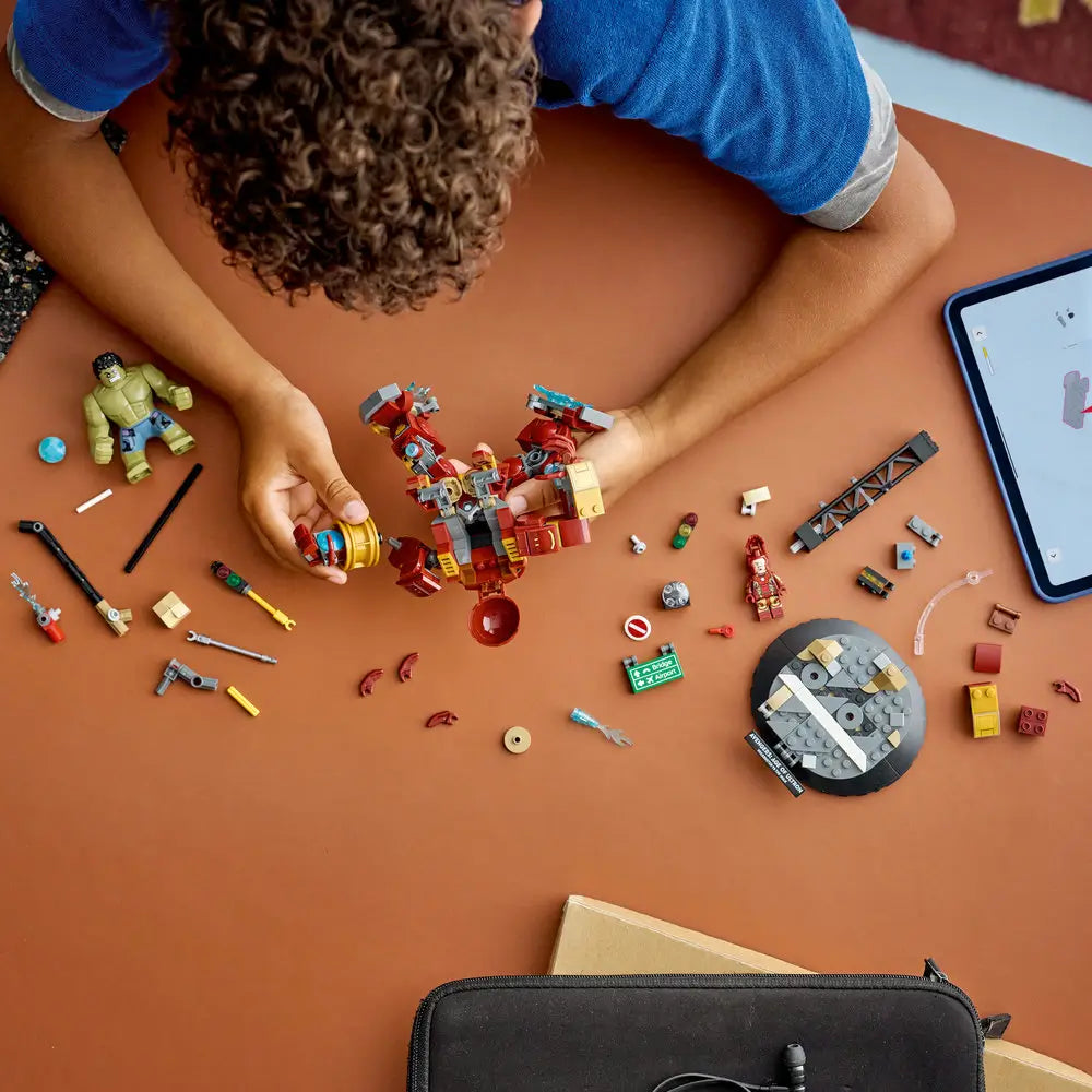 LEGO Marvel Hulkbuster build scene shows red, gold, gray, and green plastic pieces spread across a tabletop as a child assembles Hulkbuster components and accessories using step-by-step digital instructions.