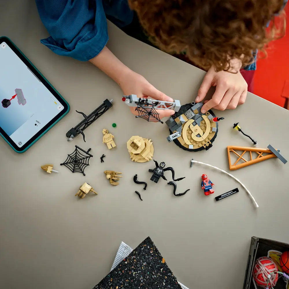LEGO Marvel building toy lifestyle image showing a child assembling the Spider-Man vs Sandman display from tan, black, and gray plastic parts on a tabletop, with web frames and character pieces laid out nearby.