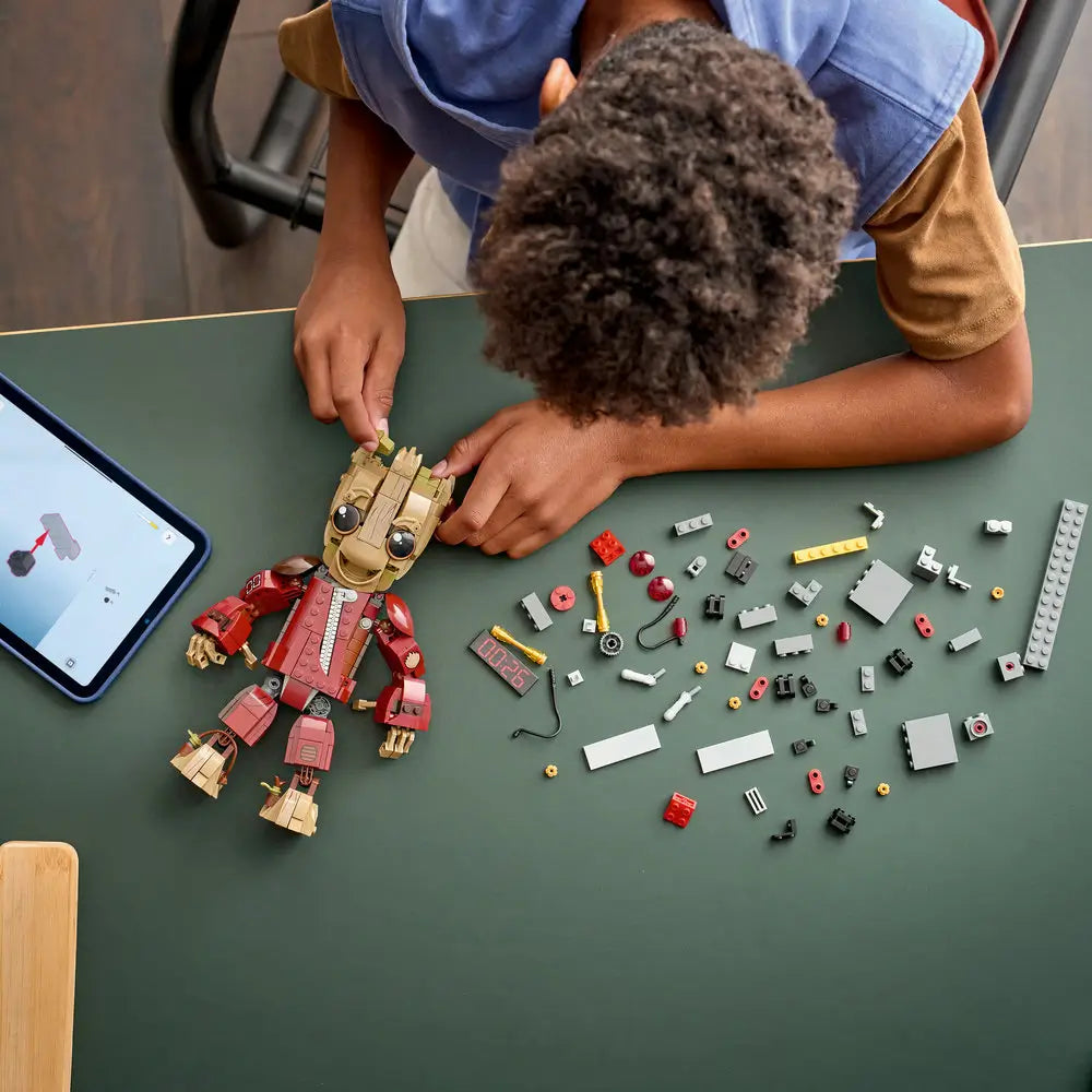 LEGO Marvel Groot lifestyle build scene shows a child assembling tan, red, and gray plastic pieces on a tabletop, following digital instructions while constructing the Groot figure and detonator accessory.