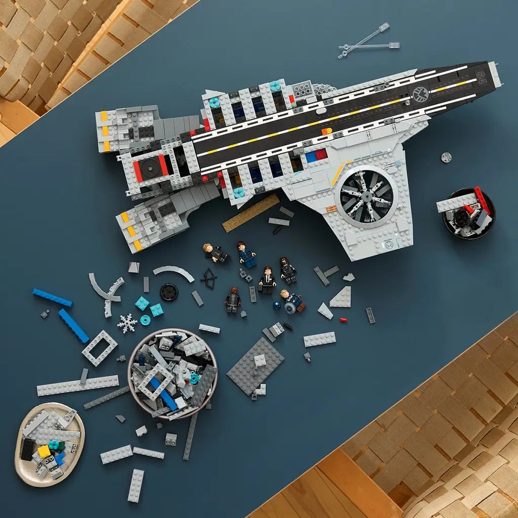 LEGO Marvel building experience, gray plastic Helicarrier sections, minifigures and loose construction pieces arranged across a tabletop during assembly of the aircraft-carrier display model.