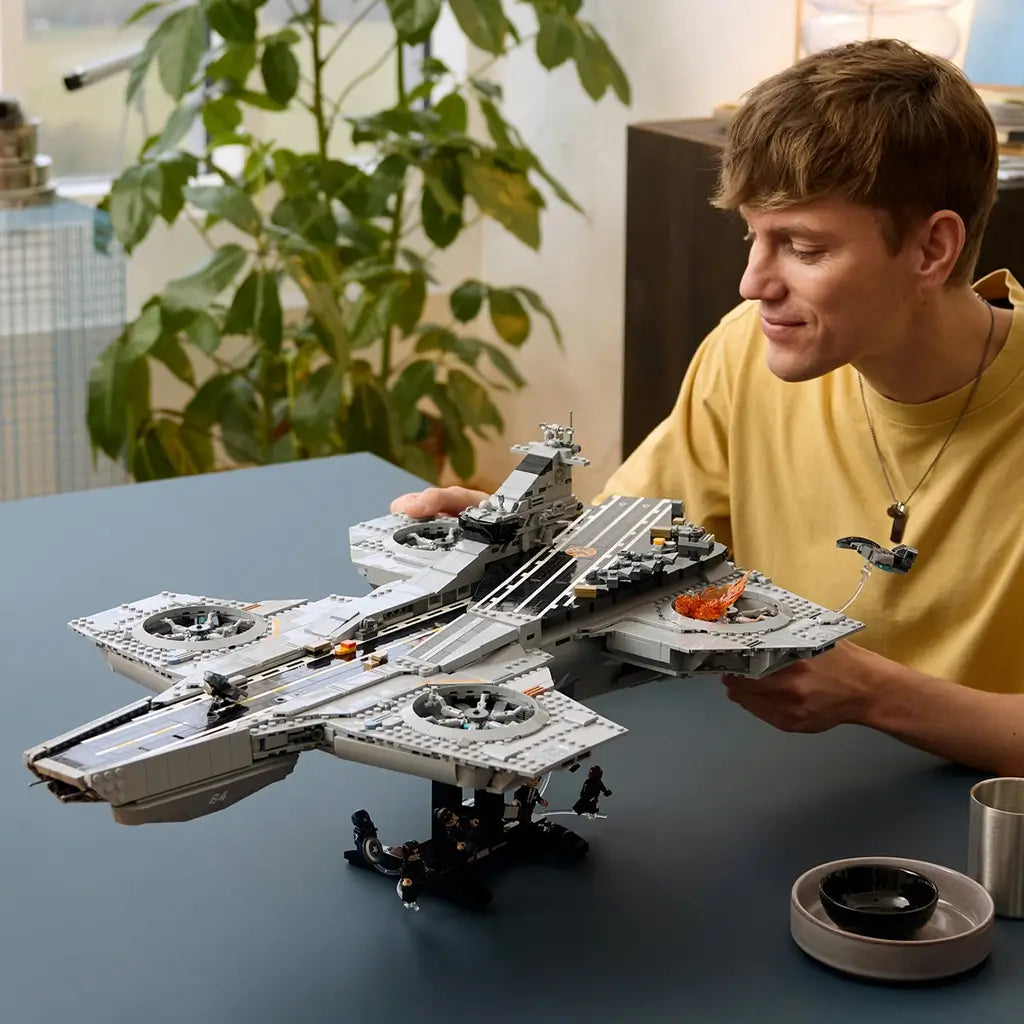 LEGO Marvel Helicarrier collectible, gray plastic aircraft-carrier model with detailed runway and rotor assemblies, presented on a stand as a builder admires it across a tabletop setting.