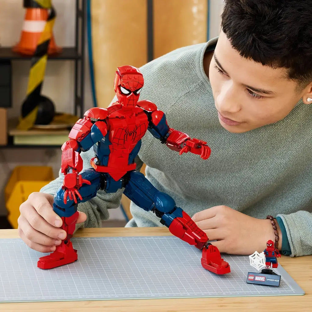 LEGO Marvel Spider-Man buildable figure, red and blue plastic superhero character with articulated joints and detailed suit markings, adjusted by a builder at a tabletop workspace.