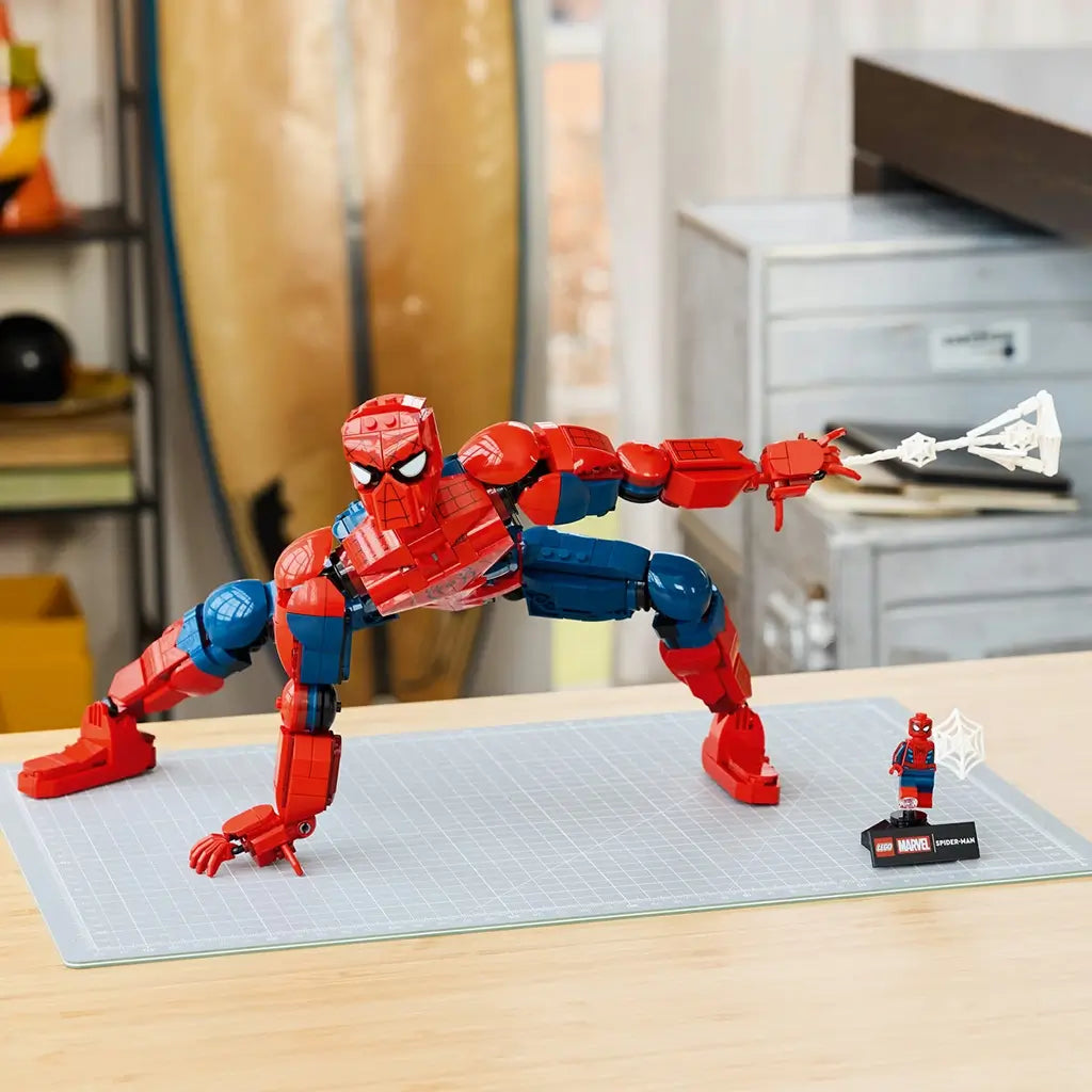 LEGO Marvel Spider-Man display model, red and blue glossy plastic superhero figure with articulated limbs and web accessory, posed on a worktable in a home setting.