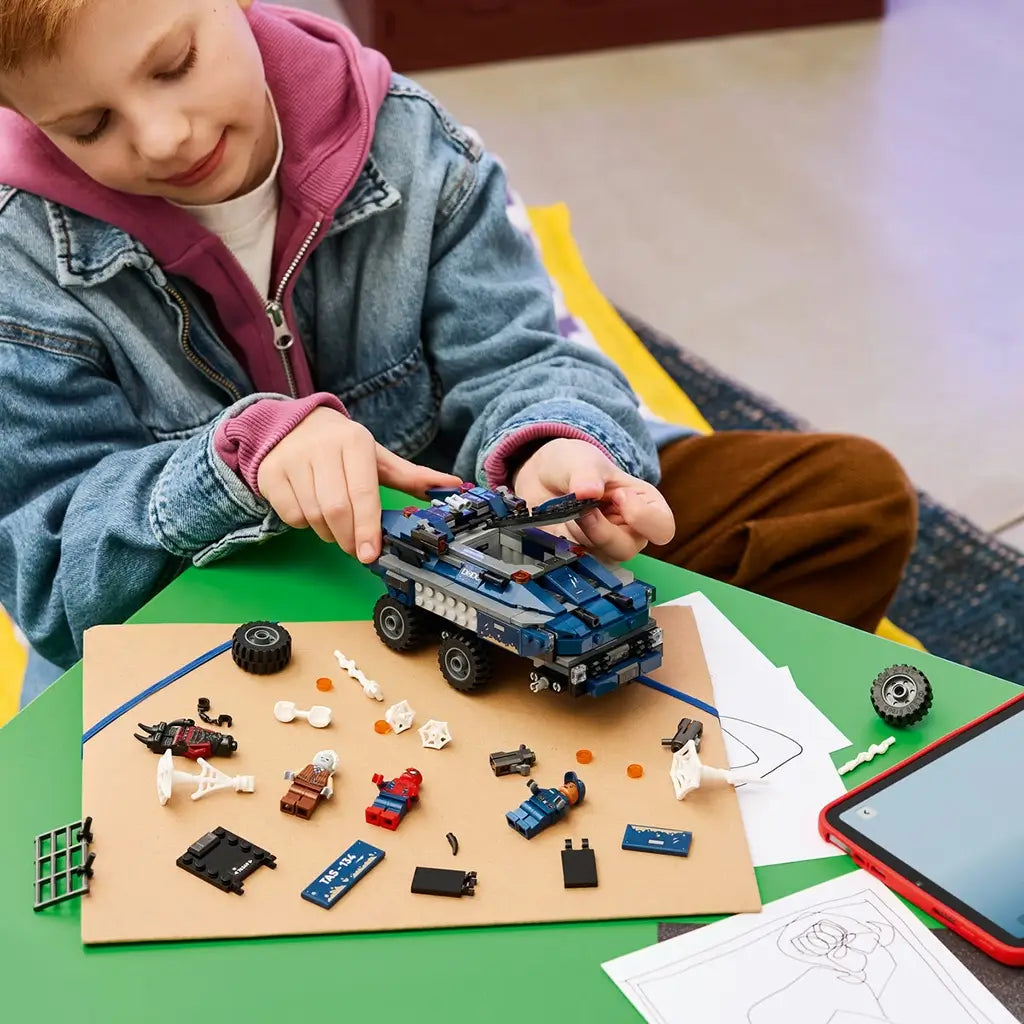 LEGO Marvel building experience, blue plastic prison transport truck components, minifigures, wheels and accessories arranged during hands-on assembly beside a tablet and drawing sheet.