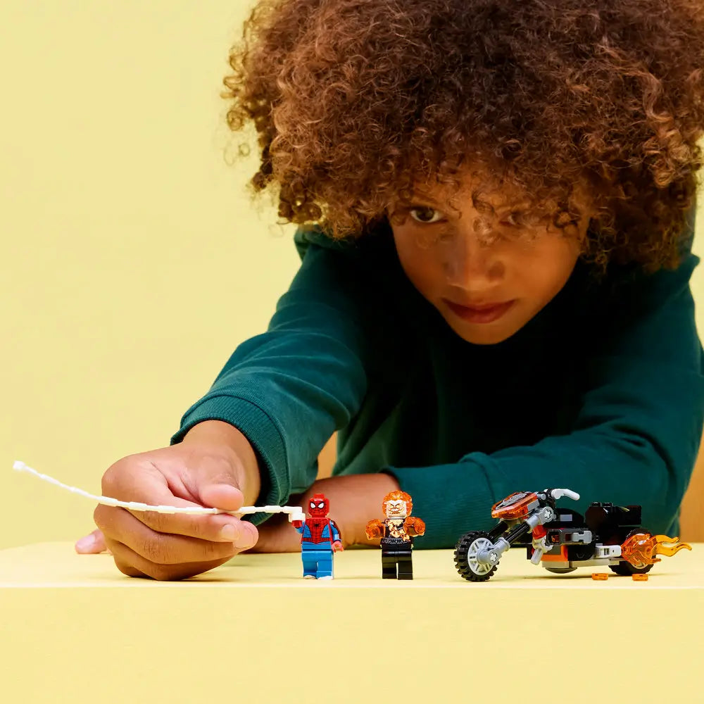 LEGO Marvel Spider-Man vs Ghost Rider display showing the black and gray motorcycle with orange flames parked beside both minifigures, photographed on a yellow studio surface to show scale, accessories, and play features.
