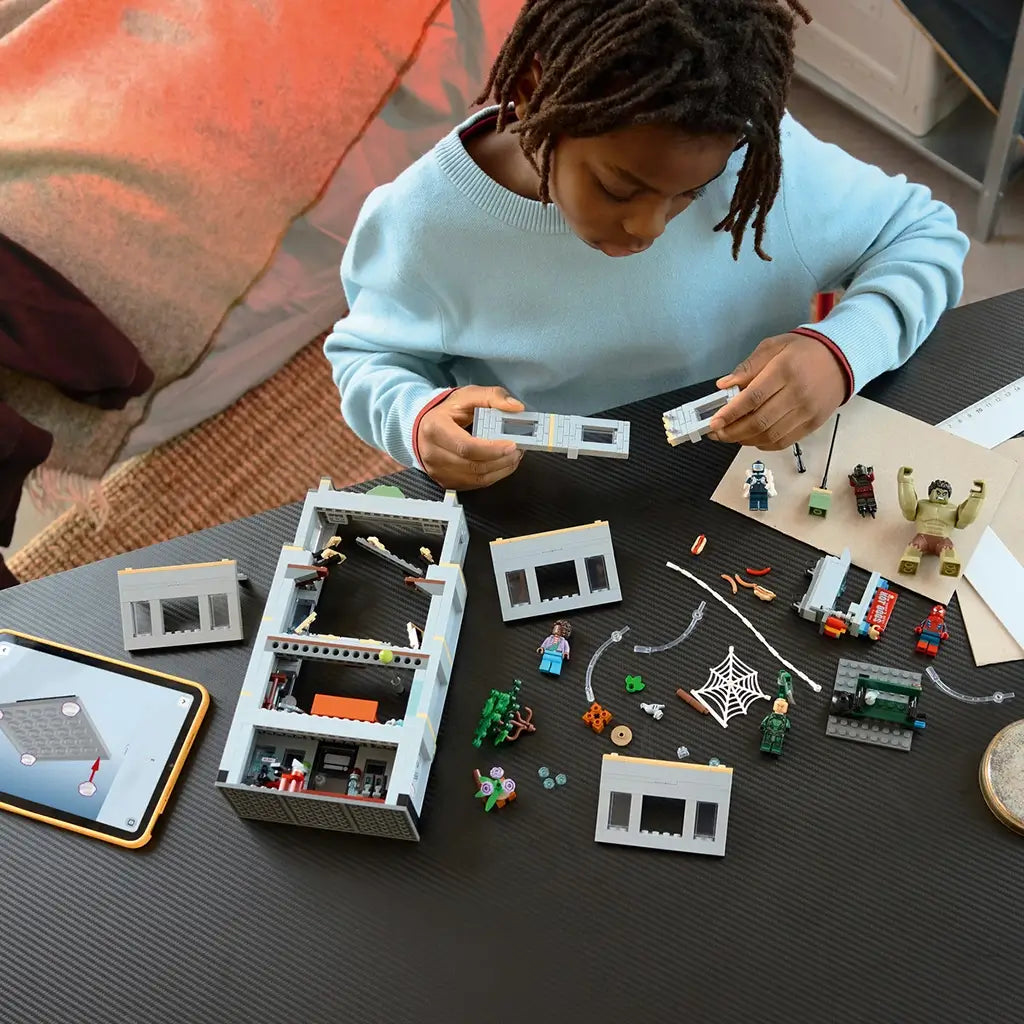 LEGO Marvel building set, gray plastic modular structure with removable wall sections, Spider-Man characters, web elements and accessories arranged during hands-on assembly on a tabletop.