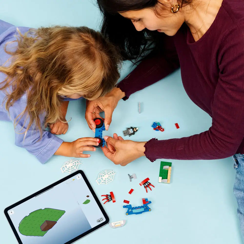 LEGO Marvel building activity scene with a child and adult assembling Spidey motorcycle pieces on a light blue table, colorful bricks, web elements, and digital instructions visible on a tablet nearby.
