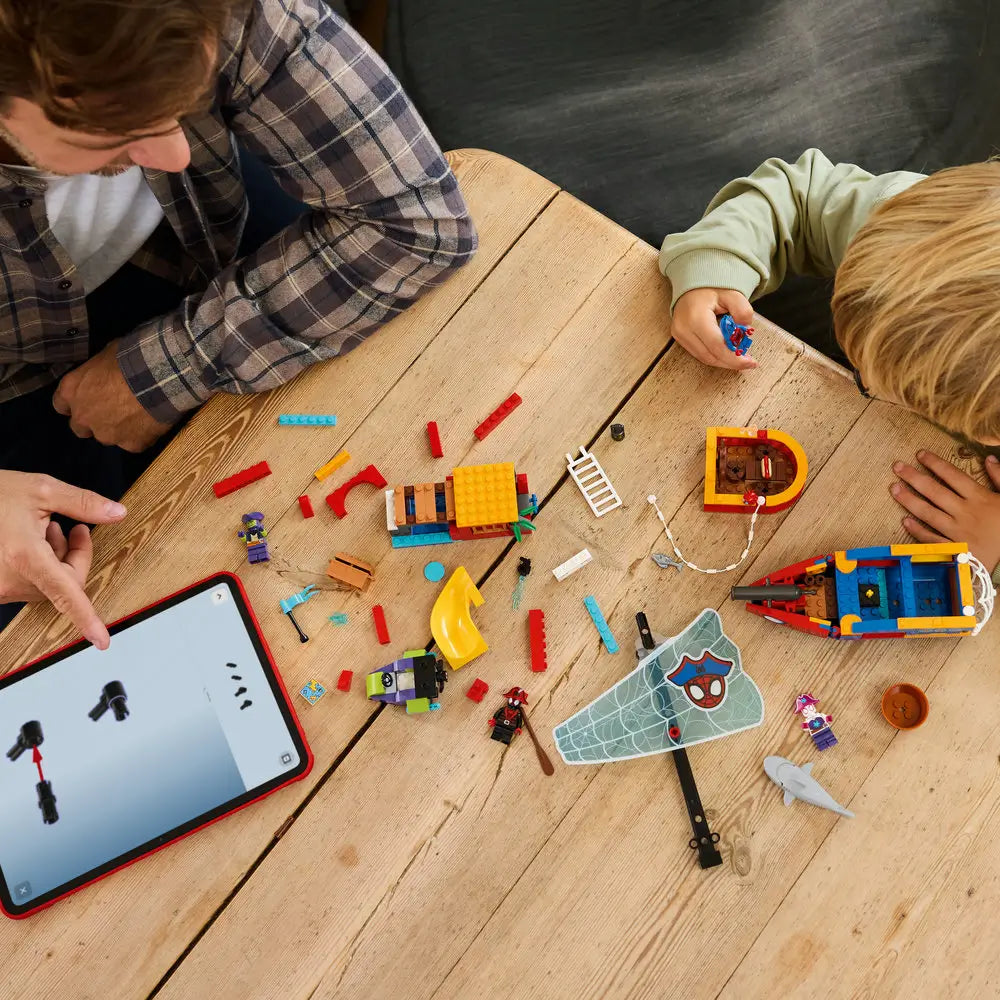 LEGO Marvel Spidey pirate ship building scene shows red, blue, and yellow plastic bricks, sail pieces, and minifigures spread across a wooden table, illustrating the guided construction process with an instruction app.