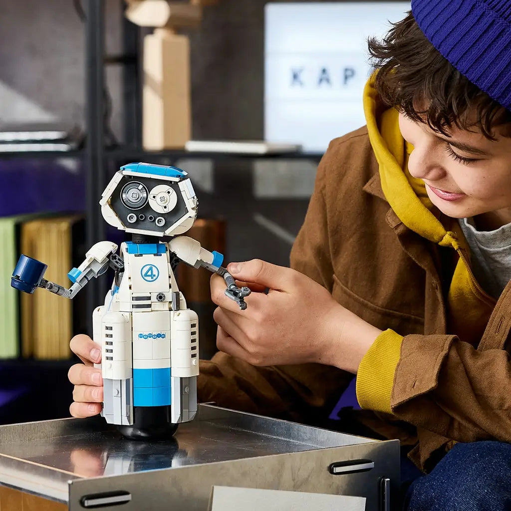 LEGO Marvel H.E.R.B.I.E. robot in white, blue and gray plastic bricks with articulated arms and rounded body, adjusted by a young builder on a metal table indoors.