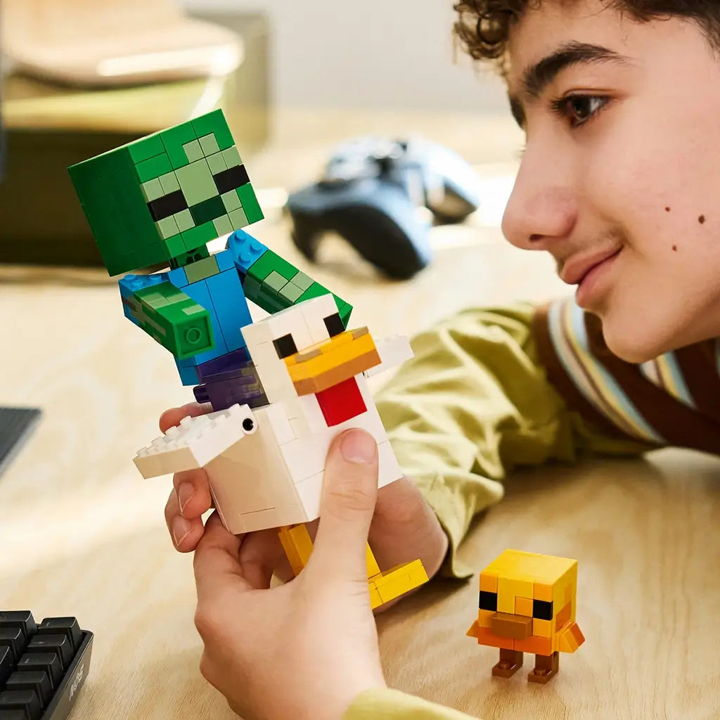 LEGO Minecraft Chicken Jockey build in white, green and yellow bricks is held by a smiling builder, with a zombie riding the large chicken and a baby chick on a wood desk.